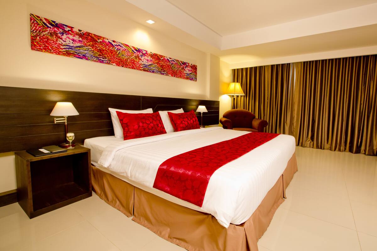 A hotel room with a large bed, a headboard, a nightstand, a lamp, a chair, a curtain, and a colorful abstract painting on the wall.