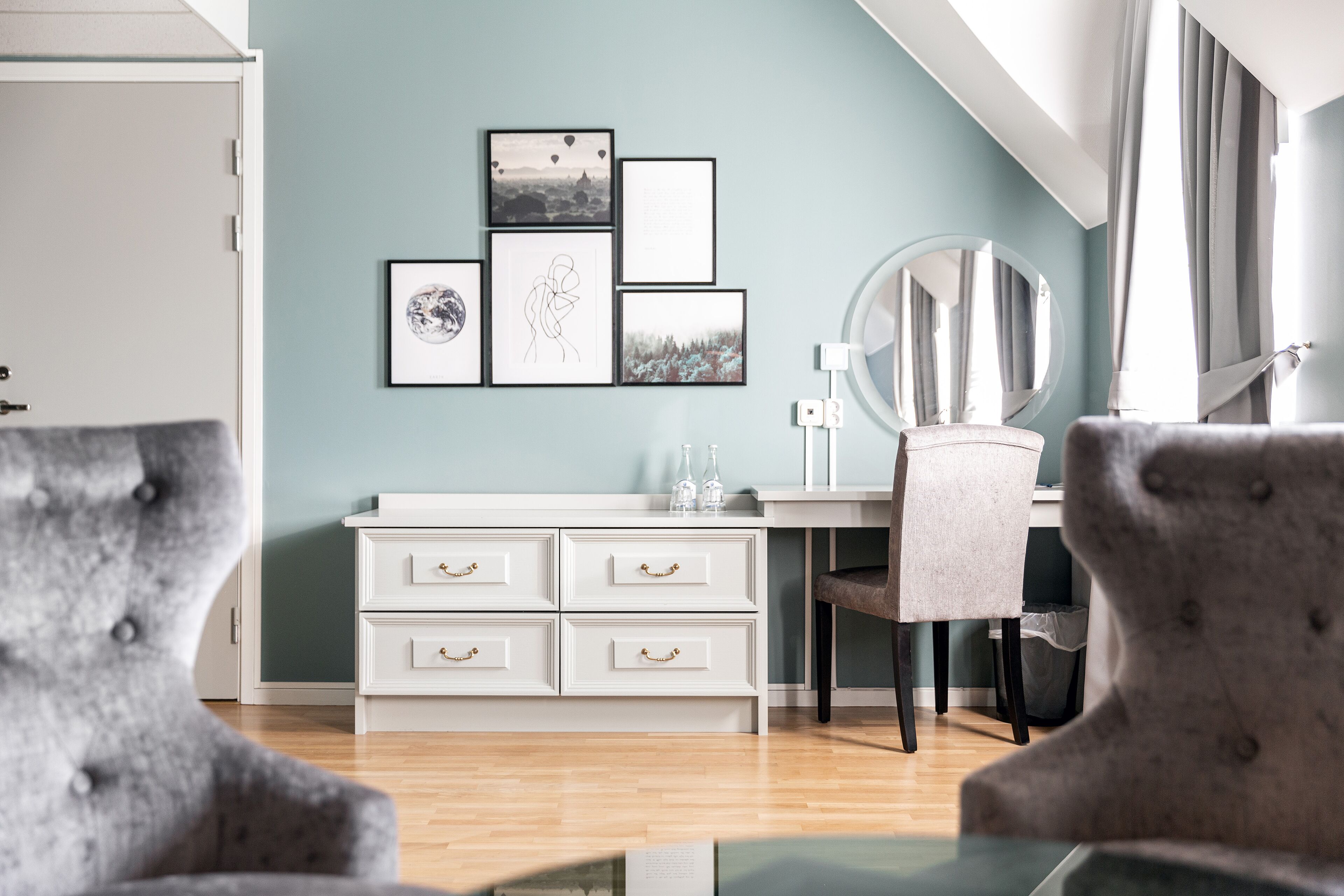A modern room with a desk, a chair, and a dresser. There are framed pictures on the wall and a round mirror above the dresser.