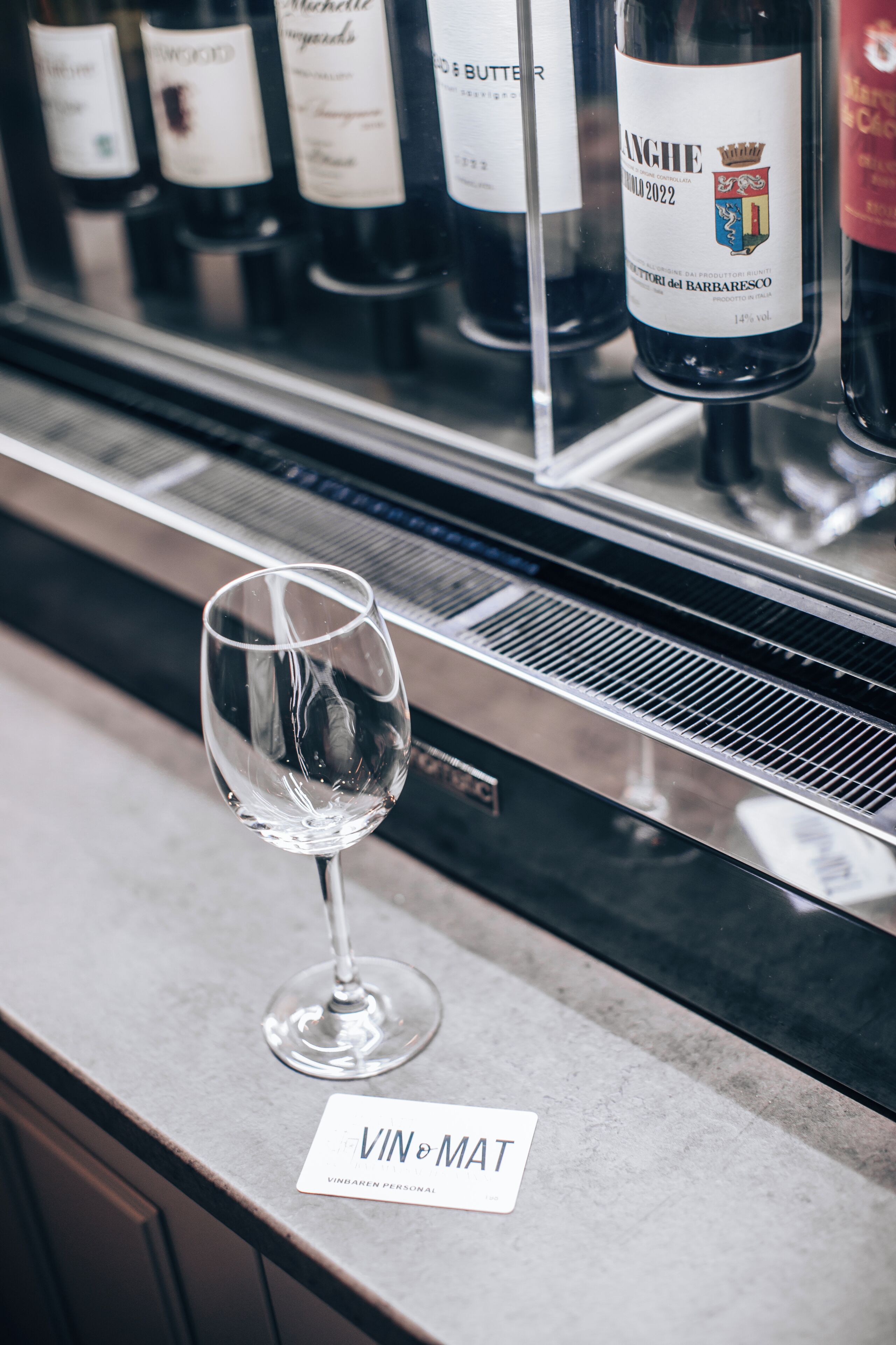 A wine glass on a counter with a label reading "VIN&MAT" and bottles of wine behind it.