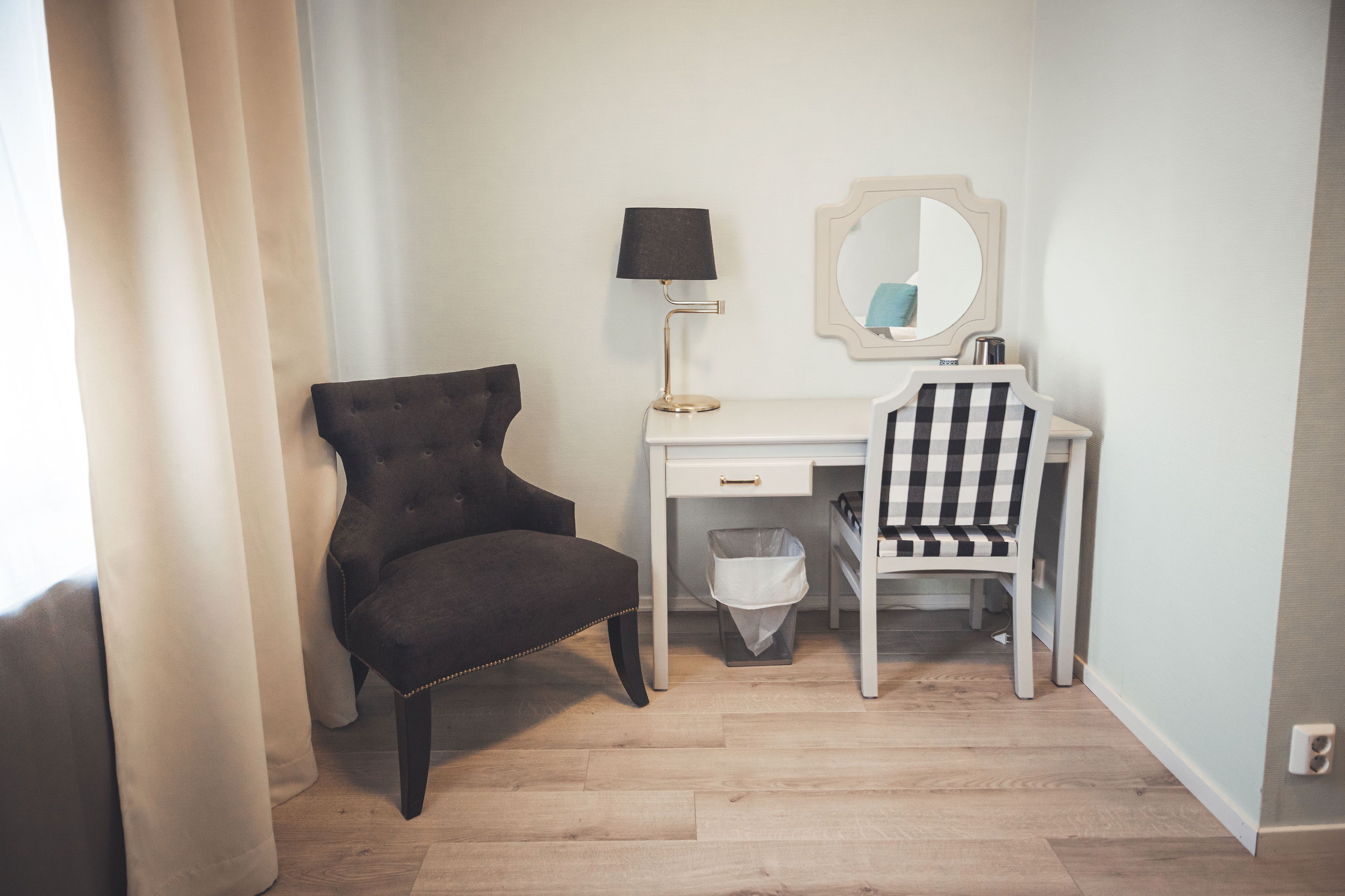 A room with a dark upholstered chair, a white desk with a mirror, a lamp, and a trash bin.