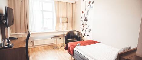 Ett hotellrum med en säng, ett skrivbord, en stol, en tv och ett fönster med gardiner.
