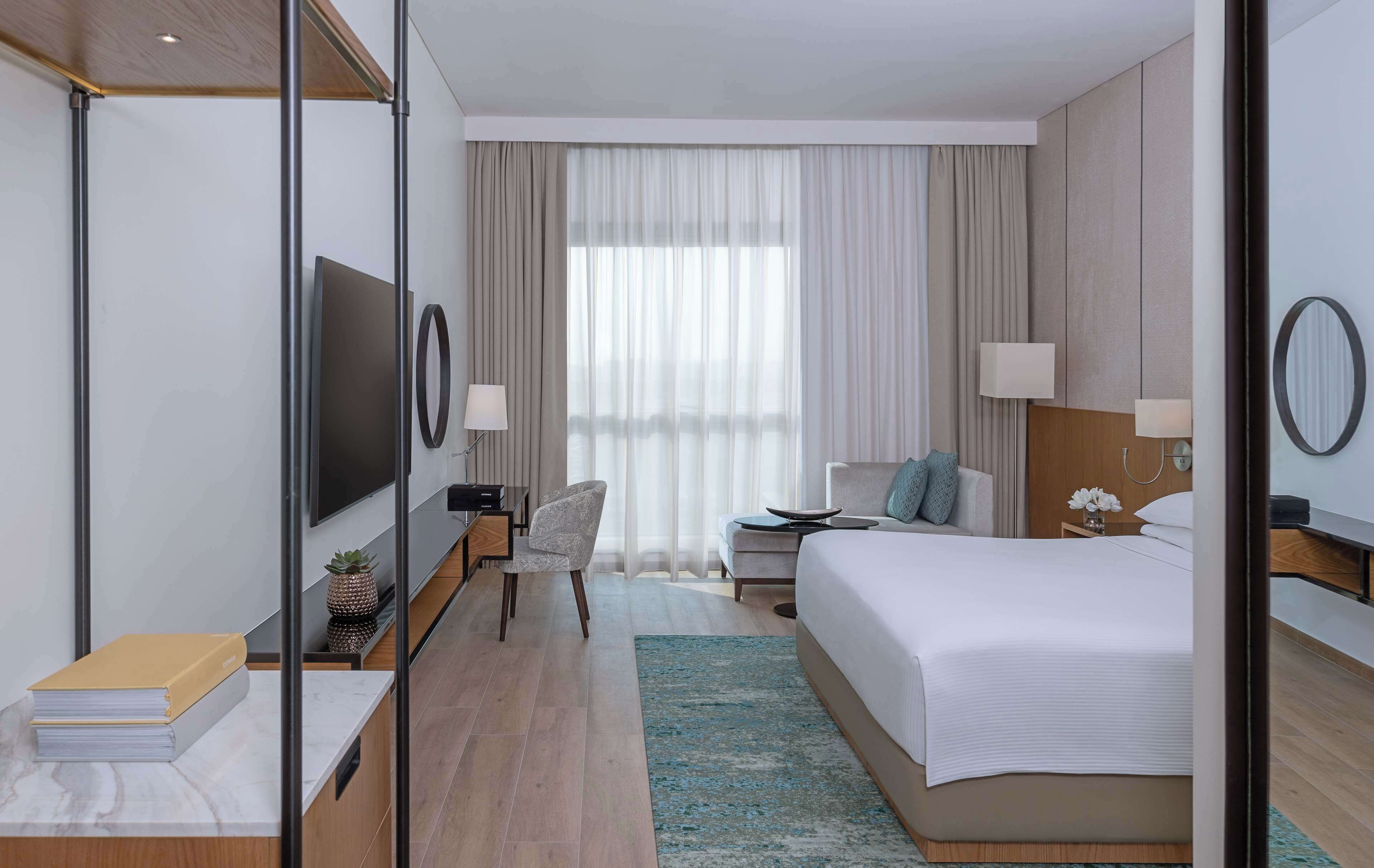 Ein modernes Hotelzimmer mit einem großen Bett, einem Schreibtisch, einem Sessel, einem Fernseher und einem Fenster mit Vorhängen.
