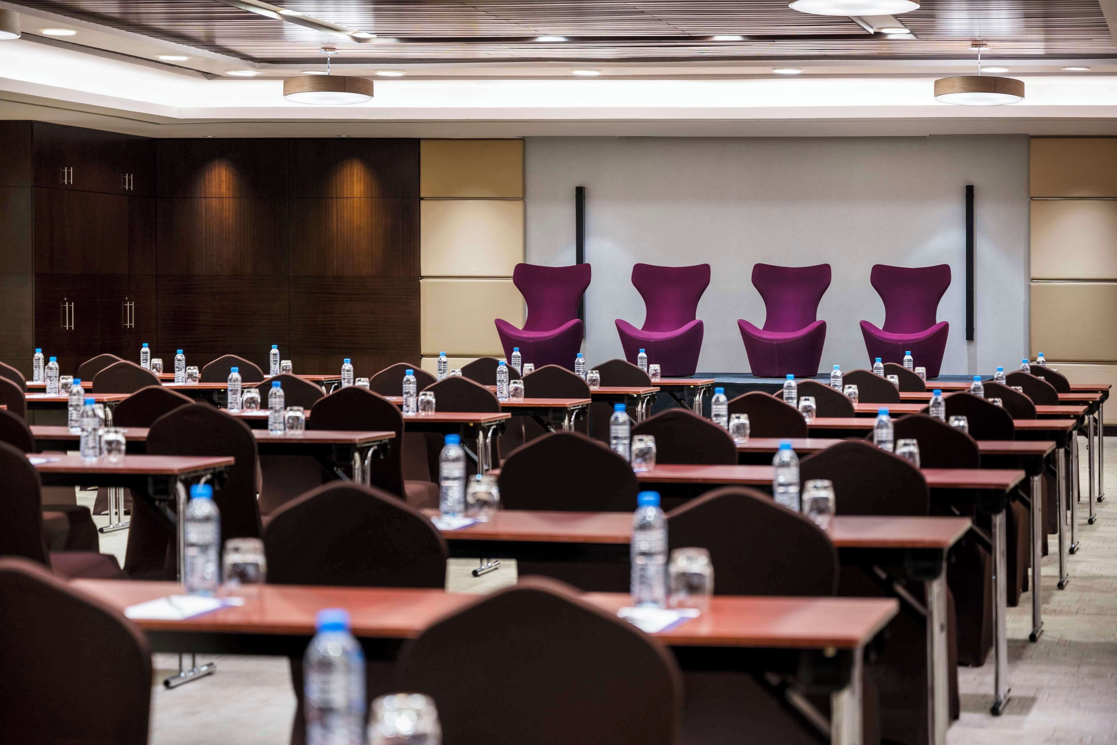 Une salle de conférence avec des rangées de tables, chacune étant équipée de bouteilles d’eau, et des chaises violettes à l’avant.