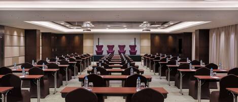 Un salón de conferencias con filas de mesas y sillas, equipado con botellas de agua y un escenario con cortinas.