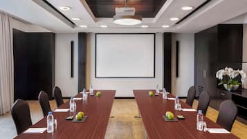 Un salón de conferencias con una larga mesa, sillas, botellas de agua, manzanas y una pantalla de proyector.