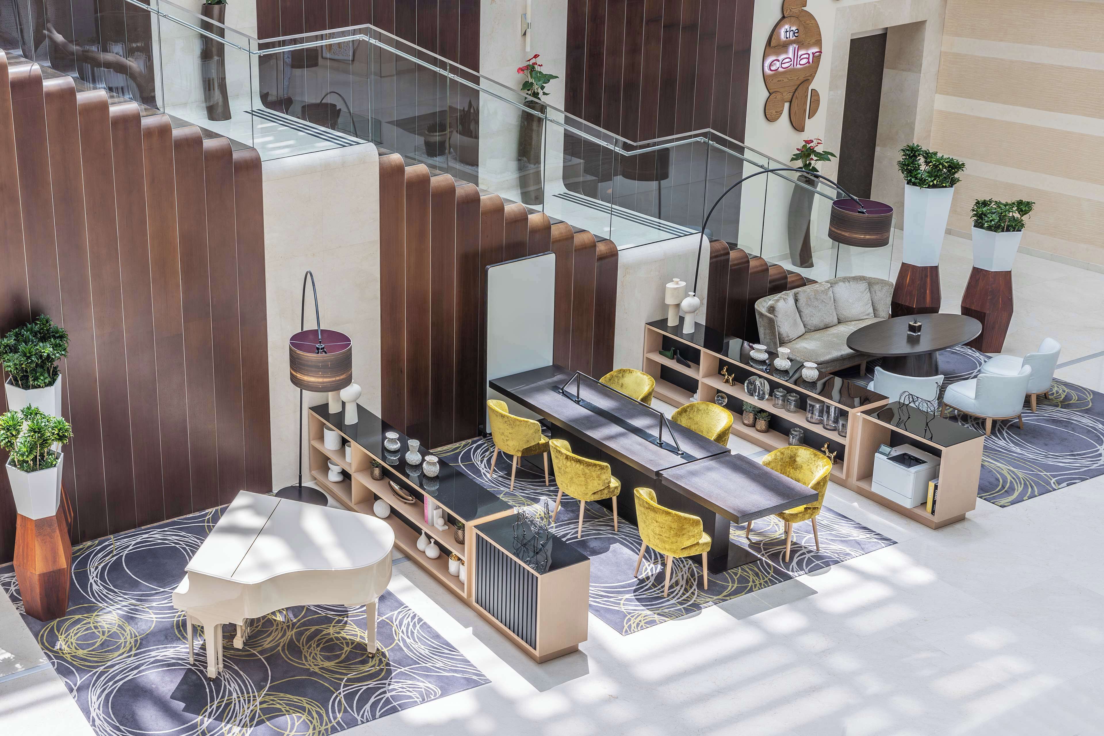 A modern lounge area with a central bar, yellow chairs, and a sofa. There are potted plants and a staircase in the background.