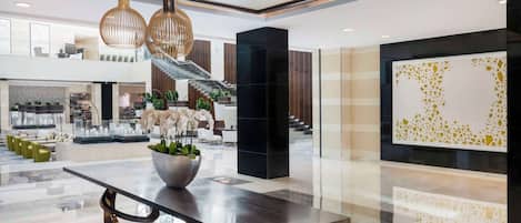 A spacious lobby with a modern design, featuring a large black reception desk, a staircase, and a decorative wall with geometric patterns.