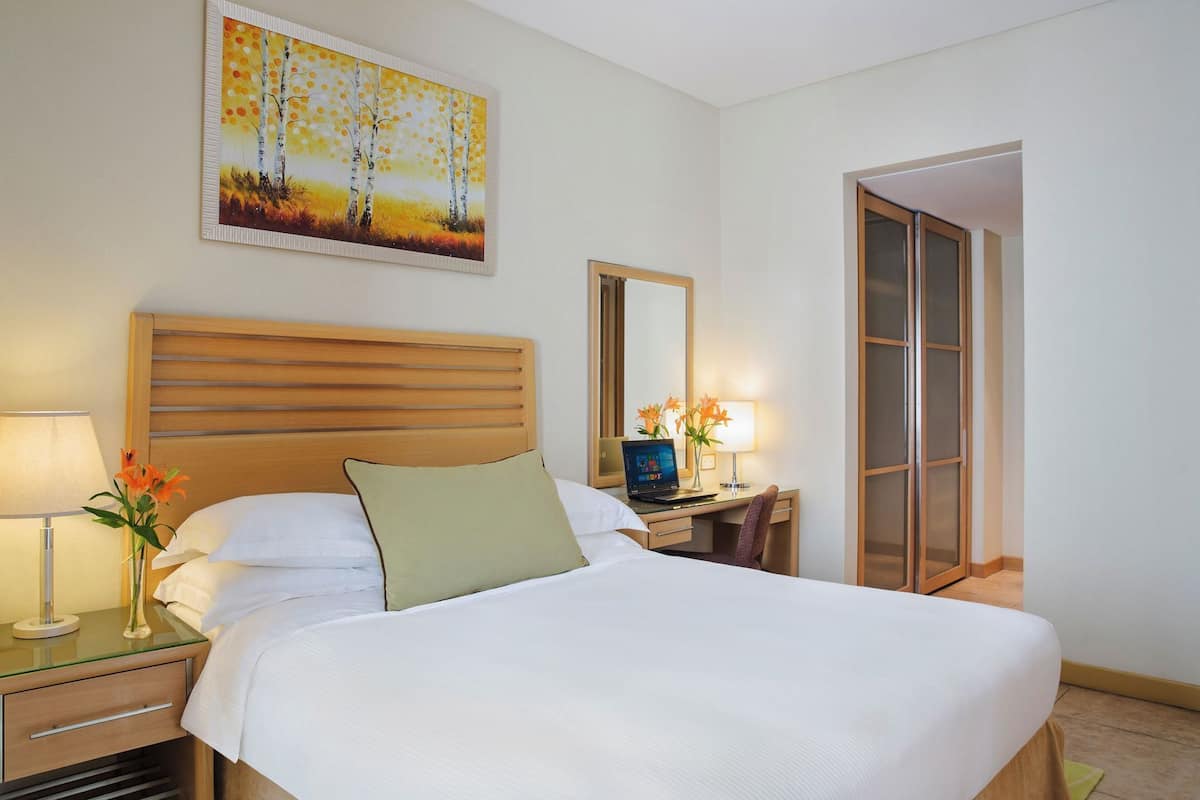 A hotel room with a large bed, a desk with a laptop, a chair, a nightstand with a lamp, and a painting on the wall.