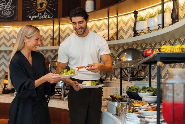 A person serving food to another person in a modern kitchen with a herringbone patterned backsplash and various kitchen appliances. - Nour Arjaan by Rotana (Al-Fujairah)