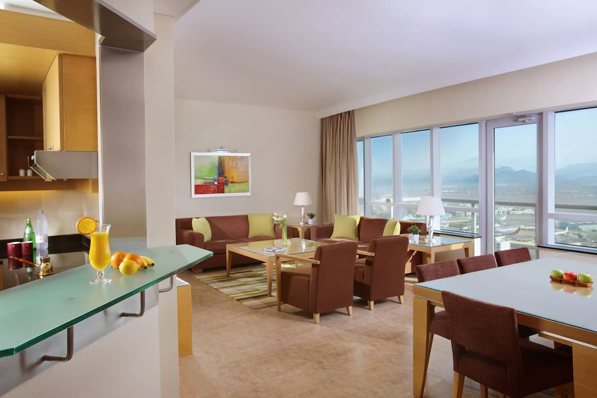 A modern hotel room with a dining area, a sofa, and a large window offering a city view.