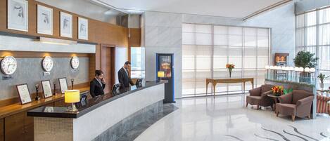 A modern hotel lobby with a reception desk, clocks, framed certificates, and a seating area with a table and chairs.