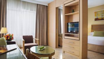 Ein Hotelzimmer mit einem Bett, einem Fernseher auf einem Holzboden, einem Esstisch mit Stühlen, einer Couch und einem kleinen Tisch mit Blumen.'