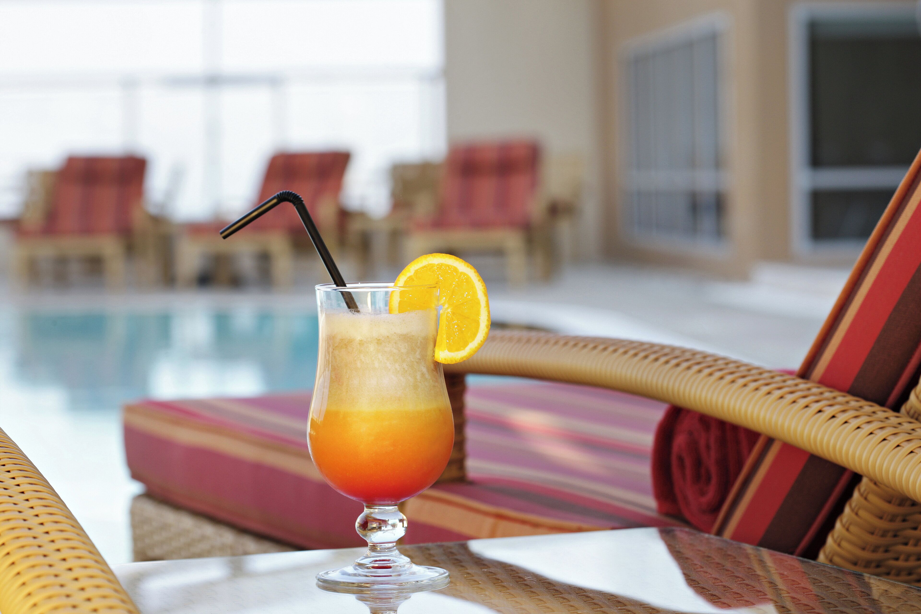 A glass of orange juice with a straw and a slice of orange on a table near a poolside lounge area.