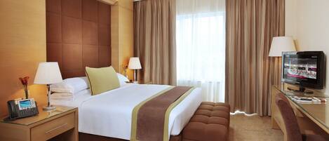A hotel room with a large bed, bedside tables, a desk with a chair, a television, and a window with curtains.