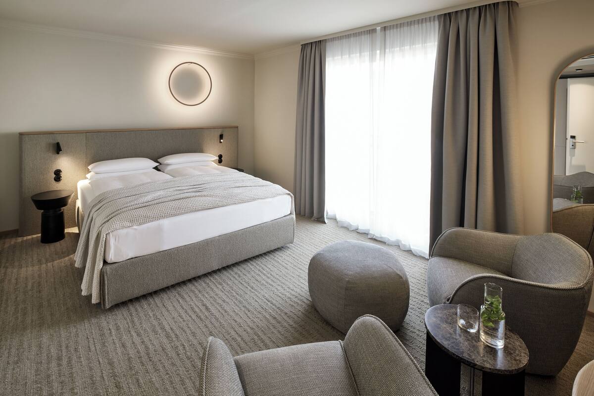 A modern hotel room with a large bed, a round wall light, a round mirror, and a seating area with a small table and chairs.