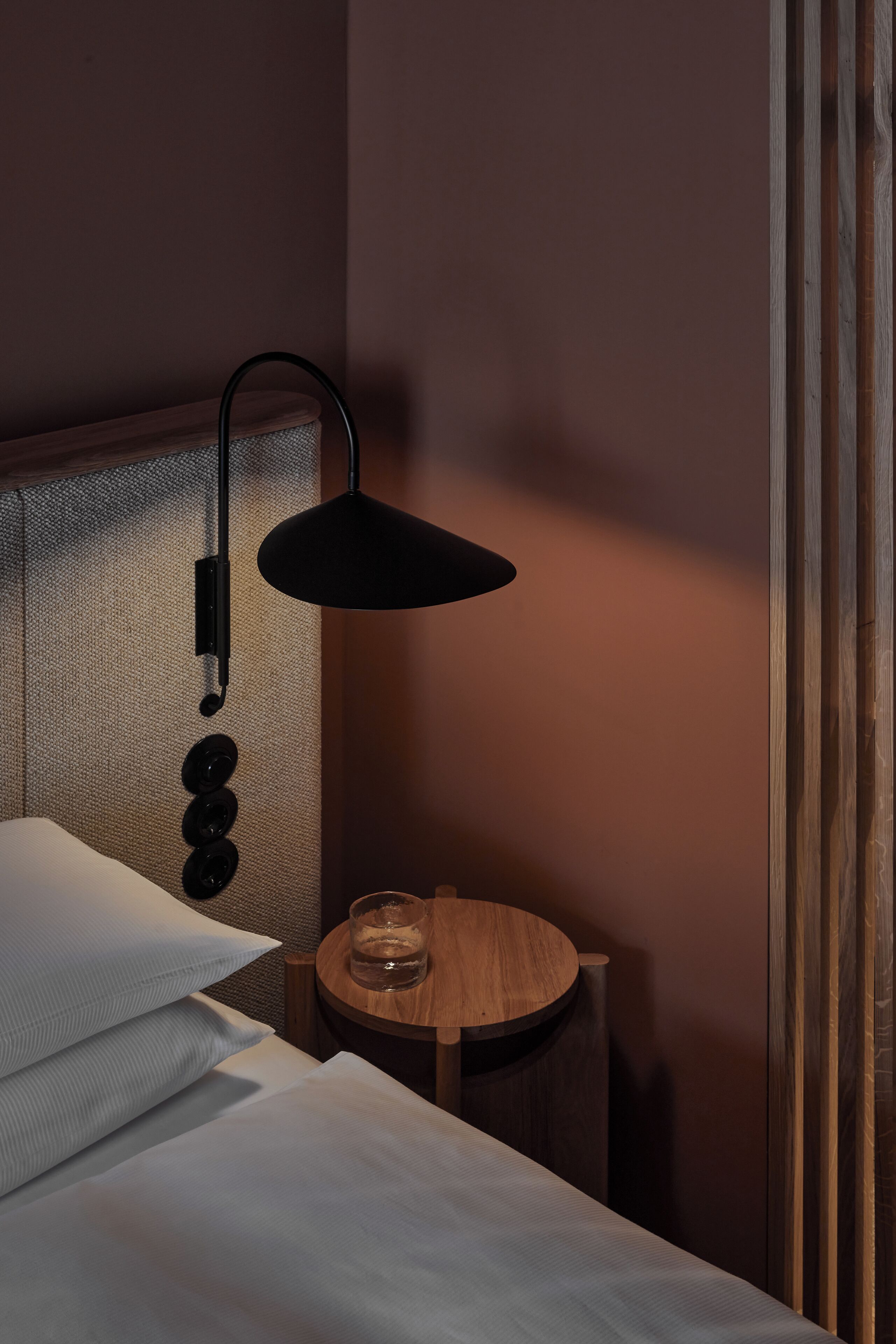 Una cama bien hecha con sábanas blancas y una mesita de noche redonda de madera. Una lámpara de escritorio negra con brazo curvado está colocada encima de la mesita.