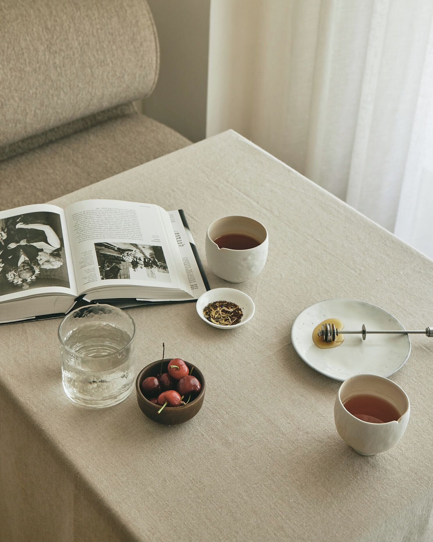 Un libro abierto sobre una mesa con una taza de té, un vaso de agua, un bol de cerezas y una cucharilla de miel.
