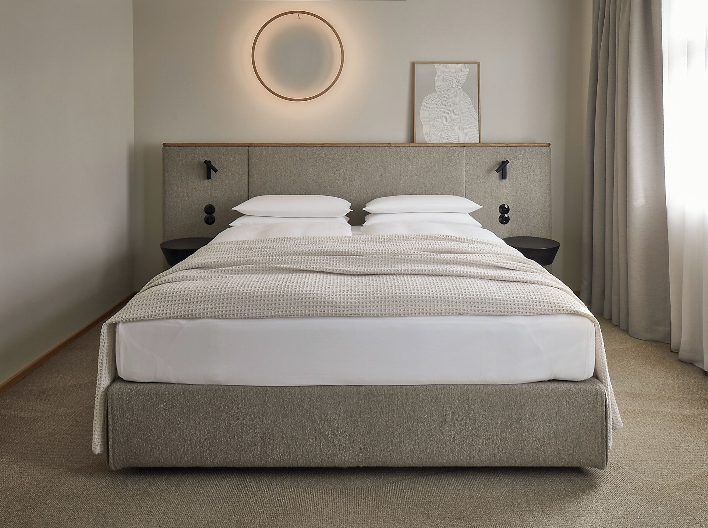 Una cama bien hecha con cabecera gris, dos mesitas de noche, una lámpara de pared redonda y un cuadro en la pared.