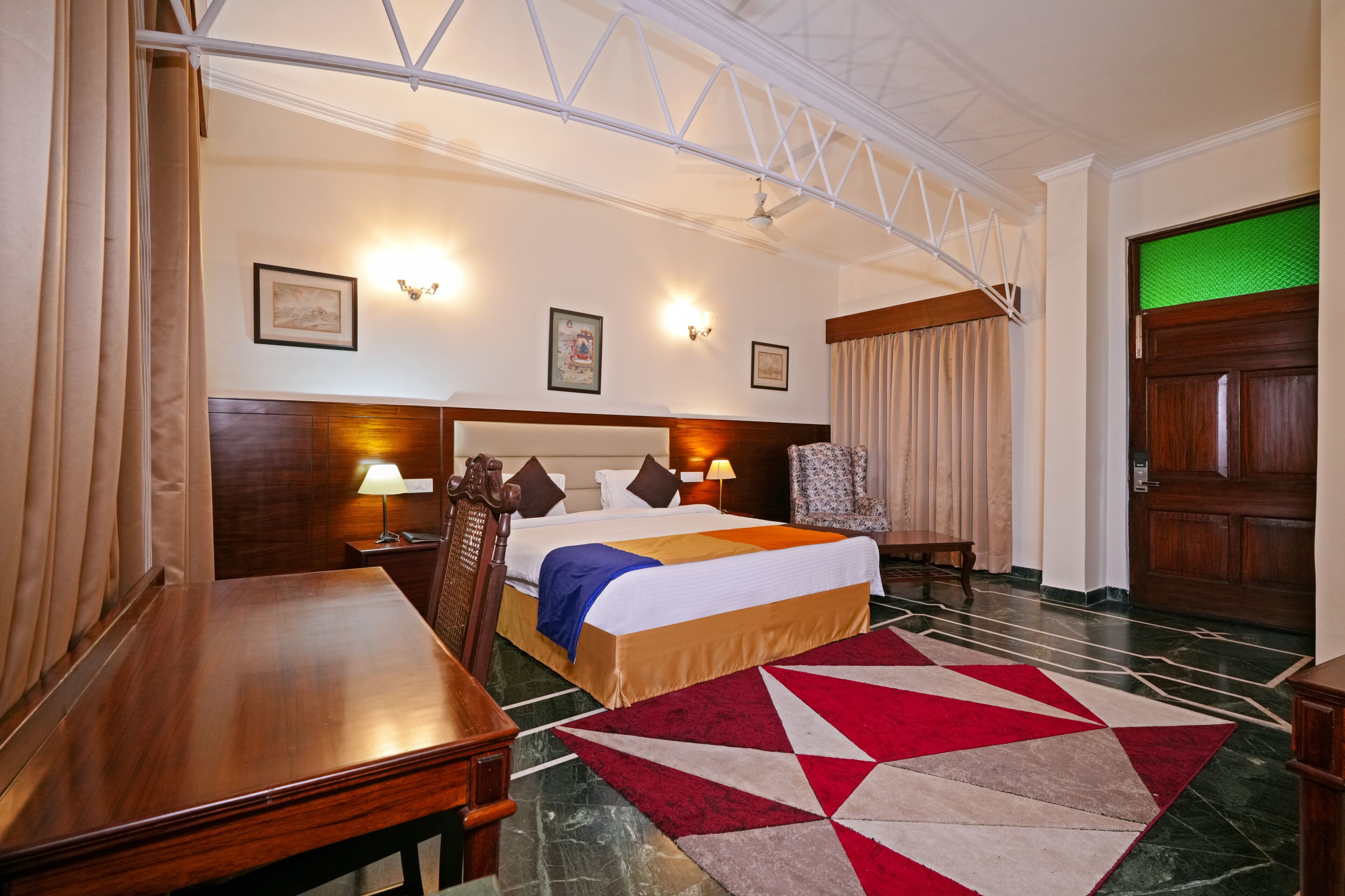 A hotel room with a large bed, a wooden desk, a chair, a red and white patterned rug, and a green door.