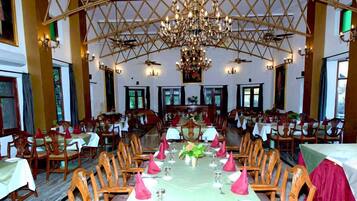 A large dining hall with multiple tables set for a meal, a chandelier, and a high ceiling with a geometric pattern.