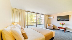 Ett hotellrum med en stor säng, en stol, ett litet bord med en vas, en platt-TV och ett fönster med gardiner.