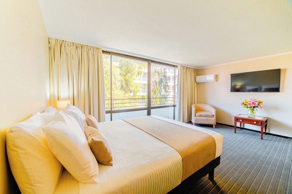 A hotel room with a large bed, a chair, a small table with a vase, a flat-screen TV, and a window with curtains.