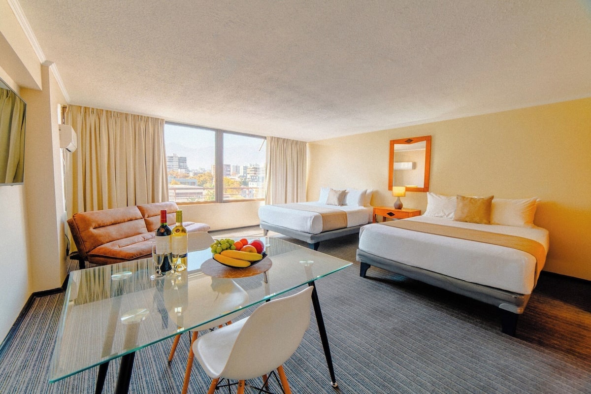 A hotel room with a glass table, two beds, a sofa, and a city view.