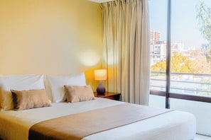 A hotel room with a large bed, a nightstand with a lamp, and a view of the city through the window. - Hotel Casa Nogales (Santiago)
