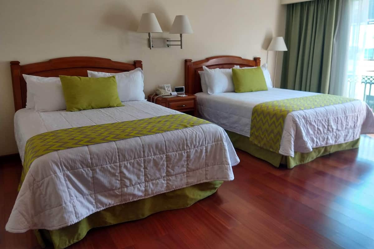 A hotel room with two beds, each with a wooden headboard, white bedding, and green accents. There are wall-mounted lamps and a window with curtains.