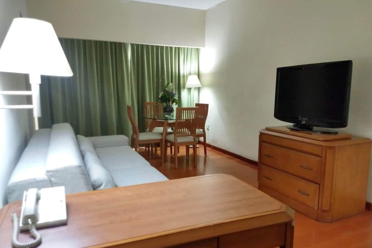 A hotel room with a sofa, a wooden table with a telephone, a television on a wooden stand, a dining table with chairs, and a green curtain.