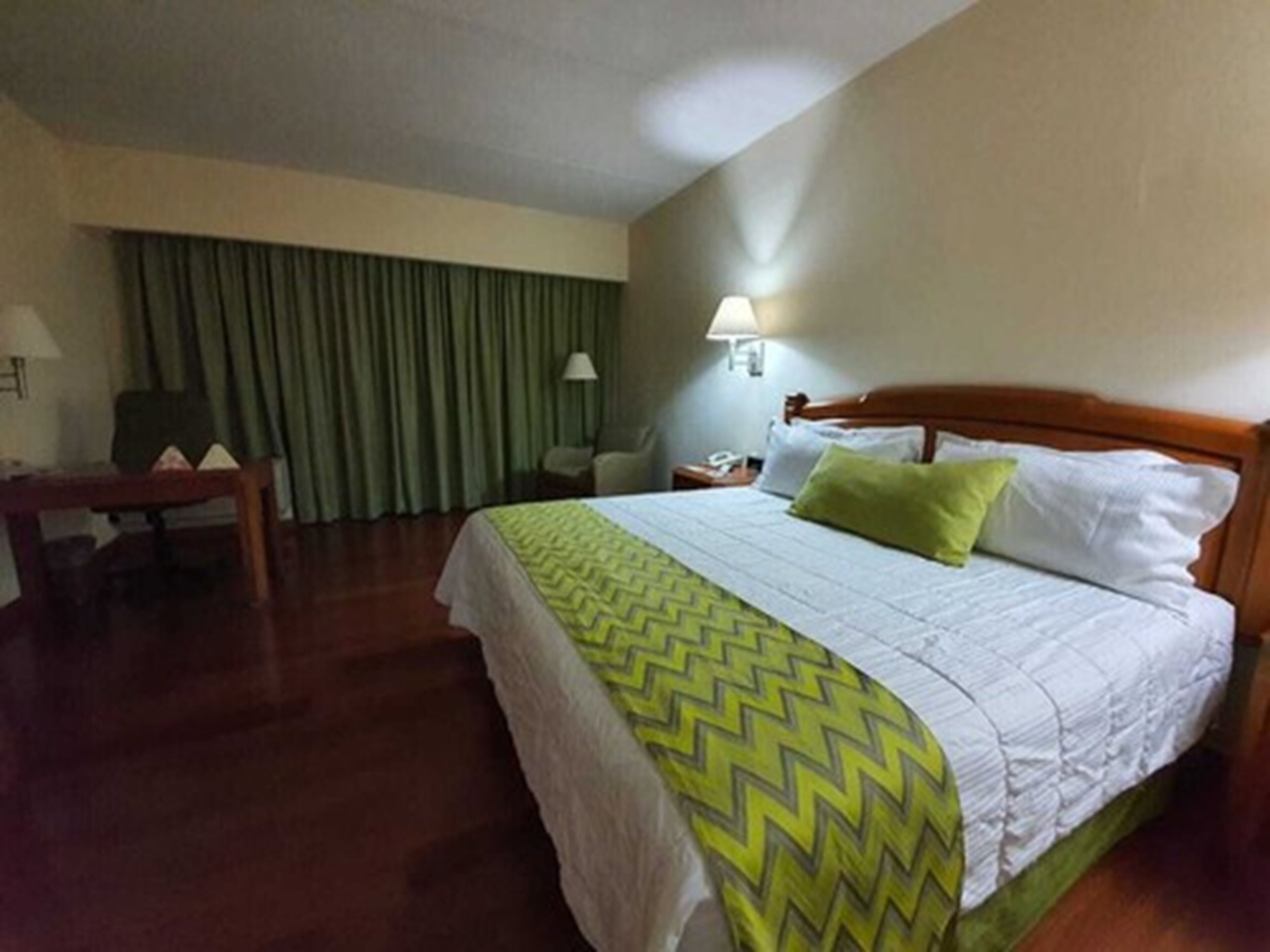 Habitació d'hotel amb llit, escriptori, cadira, làmpada i finestra amb cortines.