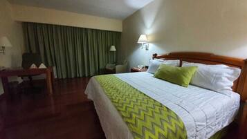 Habitació d'hotel amb llit, escriptori, cadira, làmpada i finestra amb cortines.