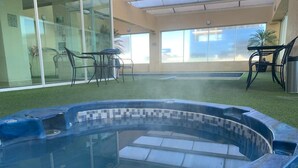 Un jacuzzi con revestimiento de azulejos, rodeado de césped artificial, y una pared de vidrio con una silla y una mesa en su interior.