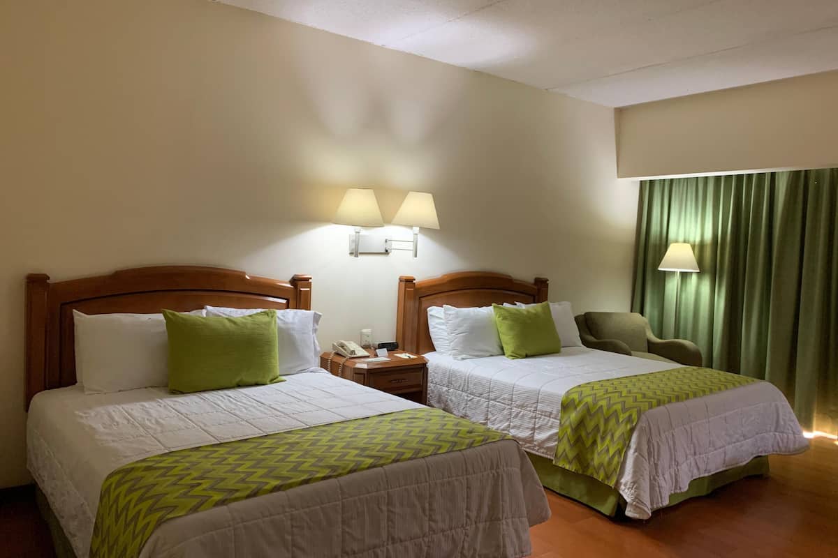 A hotel room with two beds, a chair, a desk with a phone, and a lamp.