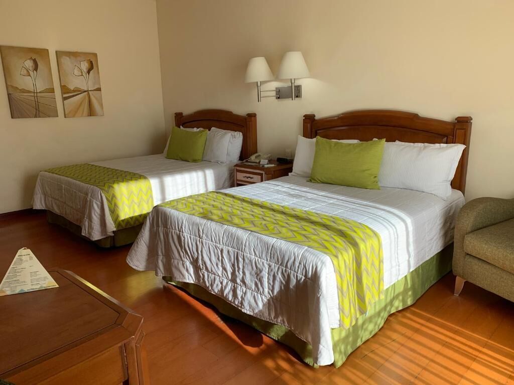 Une chambre d’hôtel avec deux lits, chacun doté d’une tête de lit en bois, de draps blancs et d’oreillers verts. Une table de chevet avec une lampe se trouve entre les deux lits, ainsi qu’une petite table sur laquelle est posé un menu.