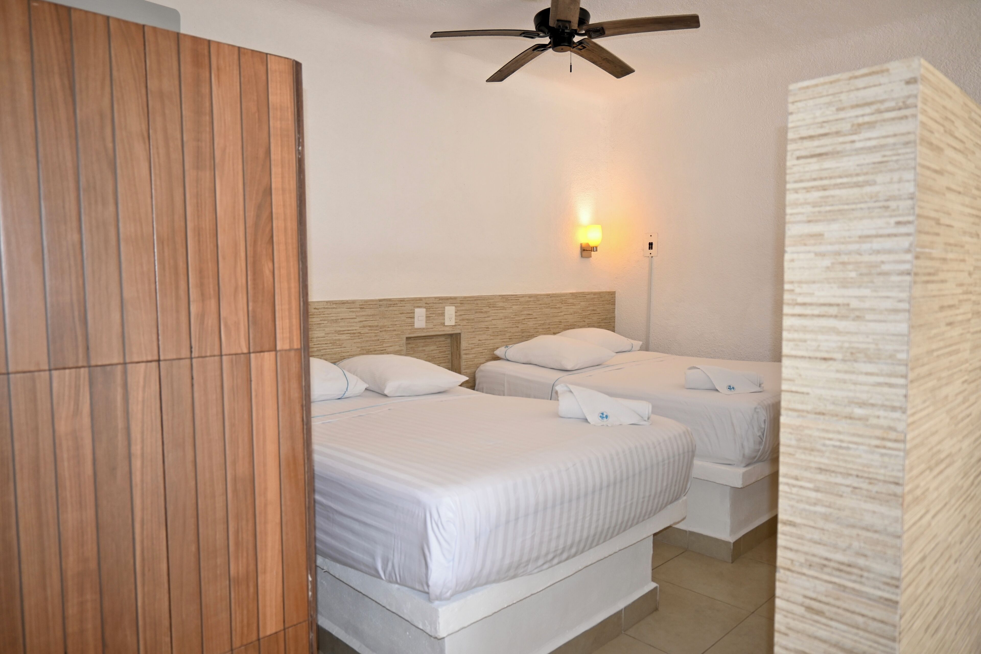 Habitación de hotel con dos camas, ventilador de techo, lámpara fijada a la pared y armario de madera.
