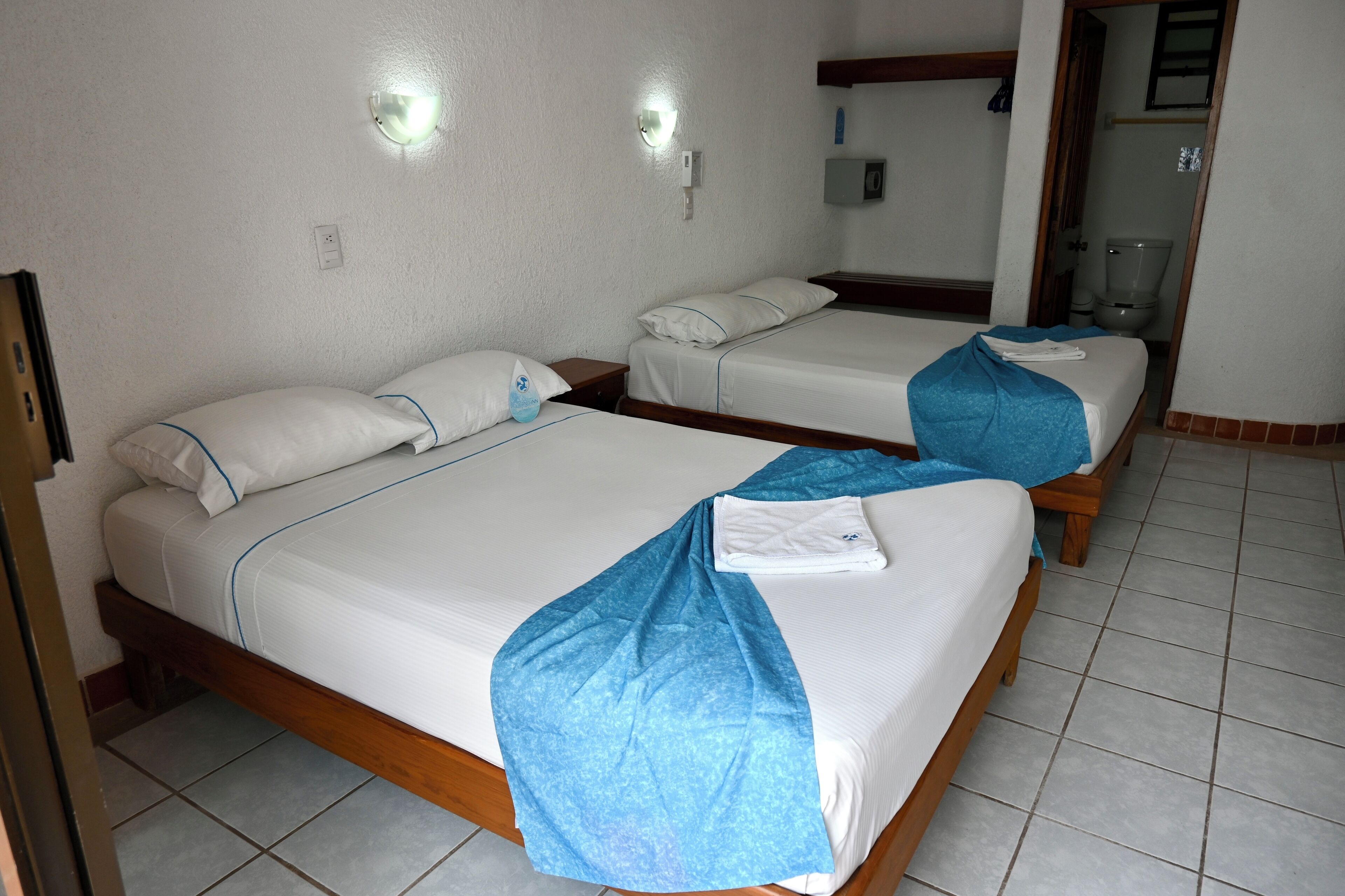 A hotel room with two single beds, each with a blue blanket and white pillows. There are wall-mounted light fixtures and a visible bathroom door.