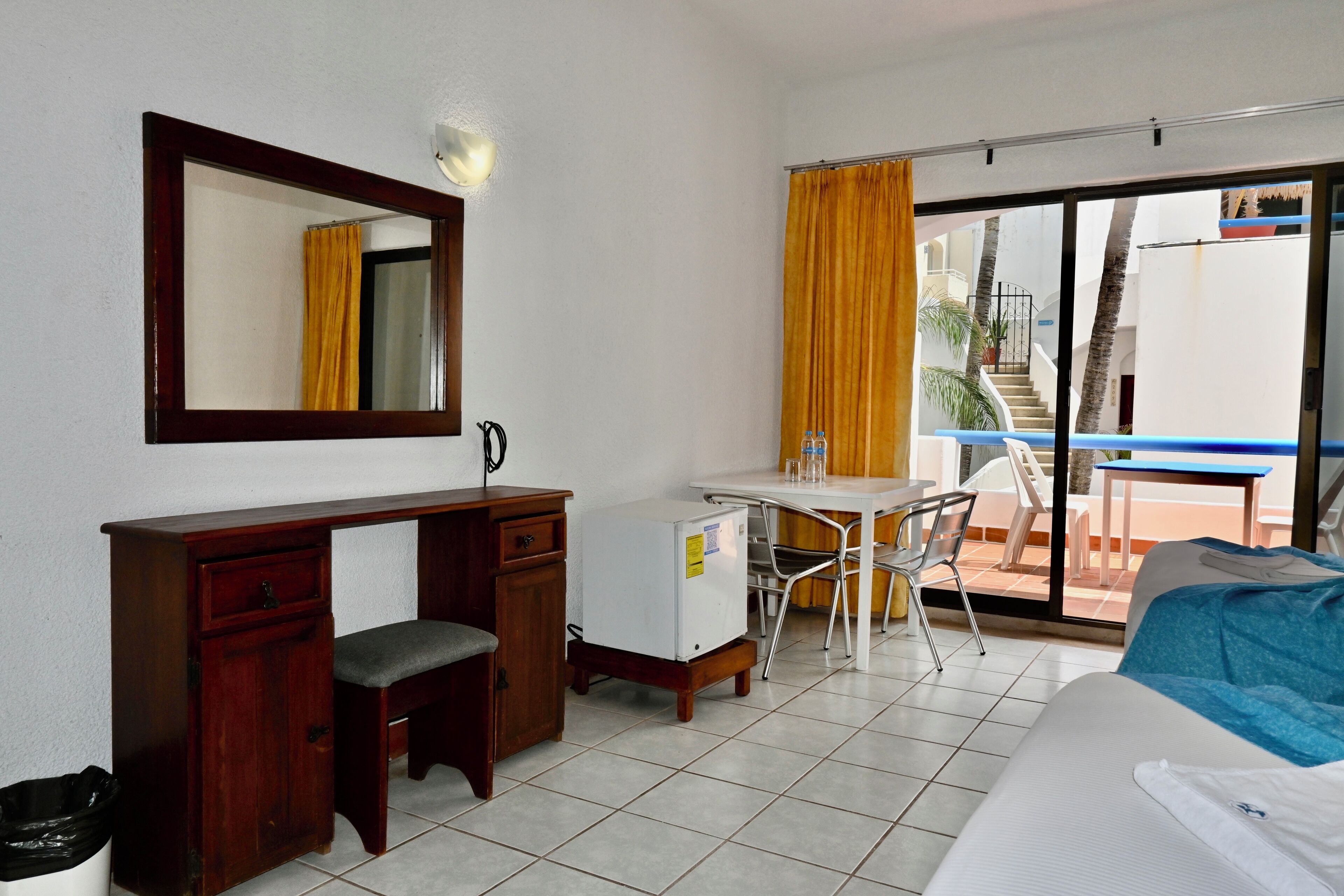 A hotel room with a desk, chair, bed, and a balcony with a view of a staircase and palm trees.