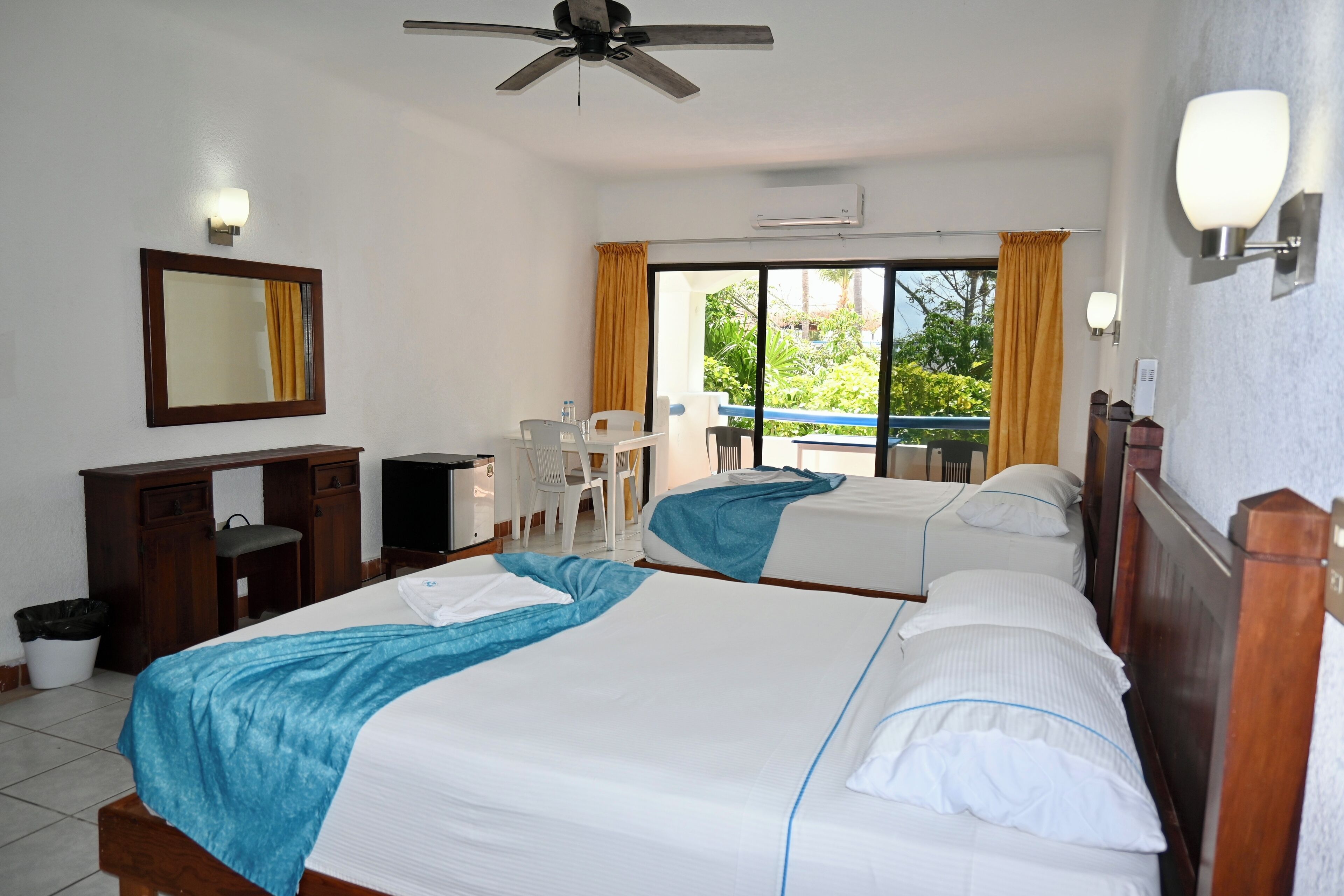 A hotel room with two beds, a desk, a chair, and a balcony with a view of greenery.