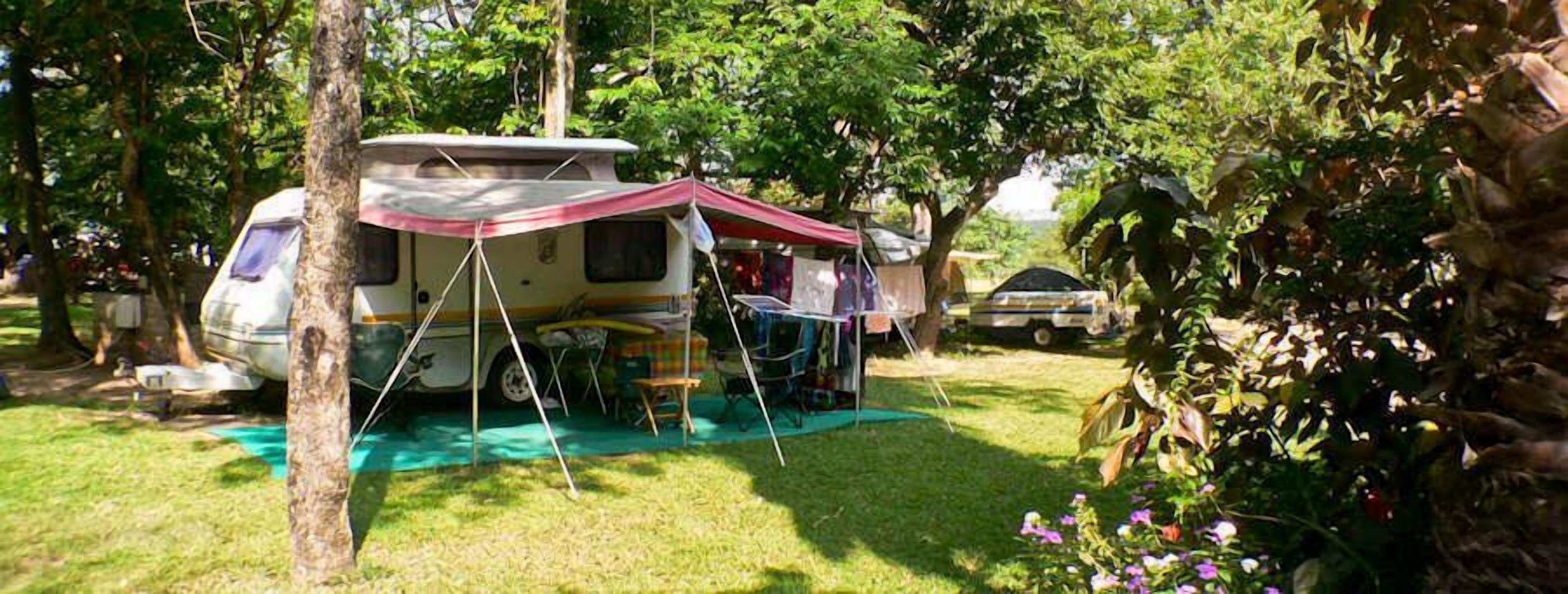 A camping site with a caravan set up under a tent, surrounded by trees and grass.