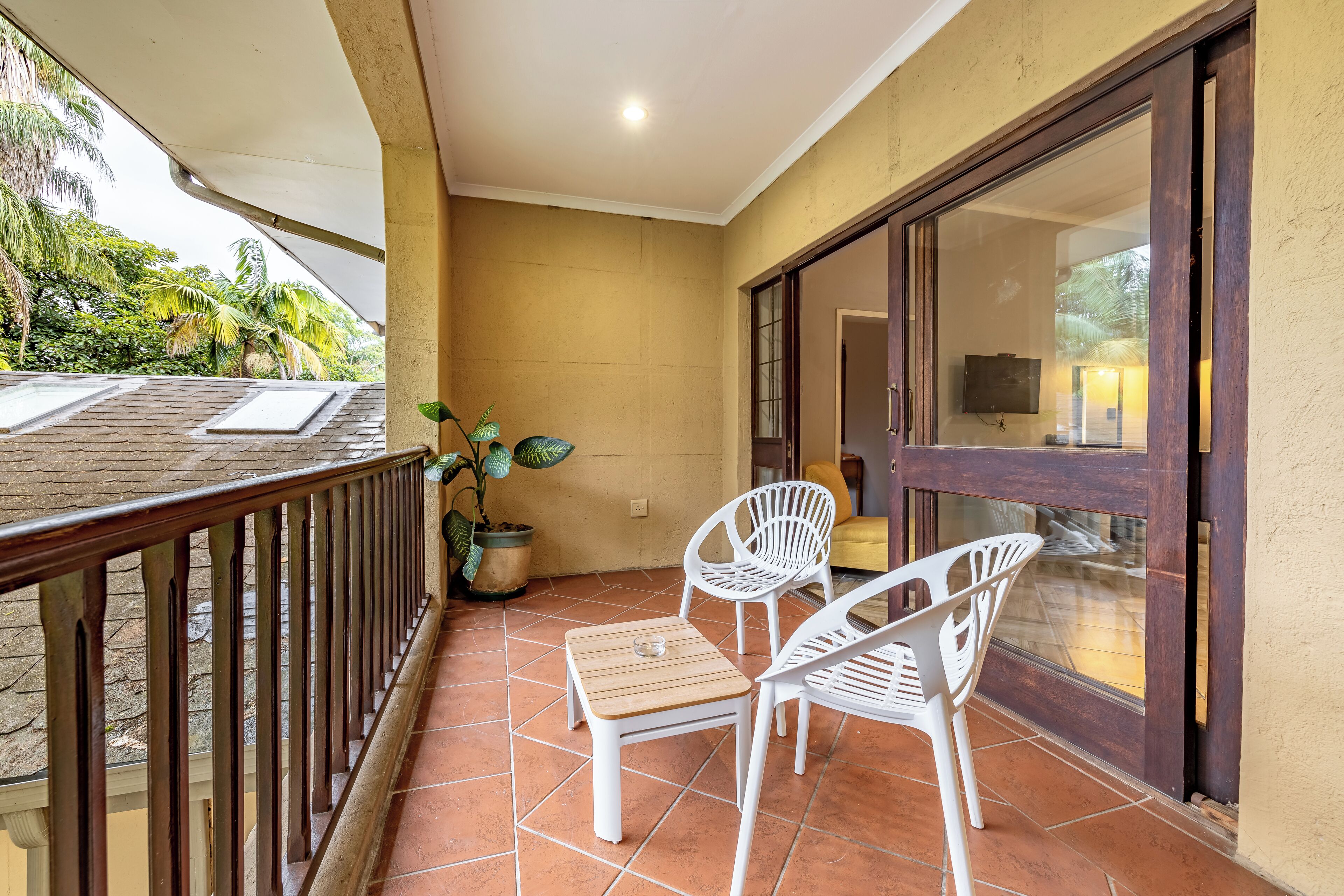 A balcony with a wooden table and two white chairs, a potted plant, and a view of the interior through a glass door.