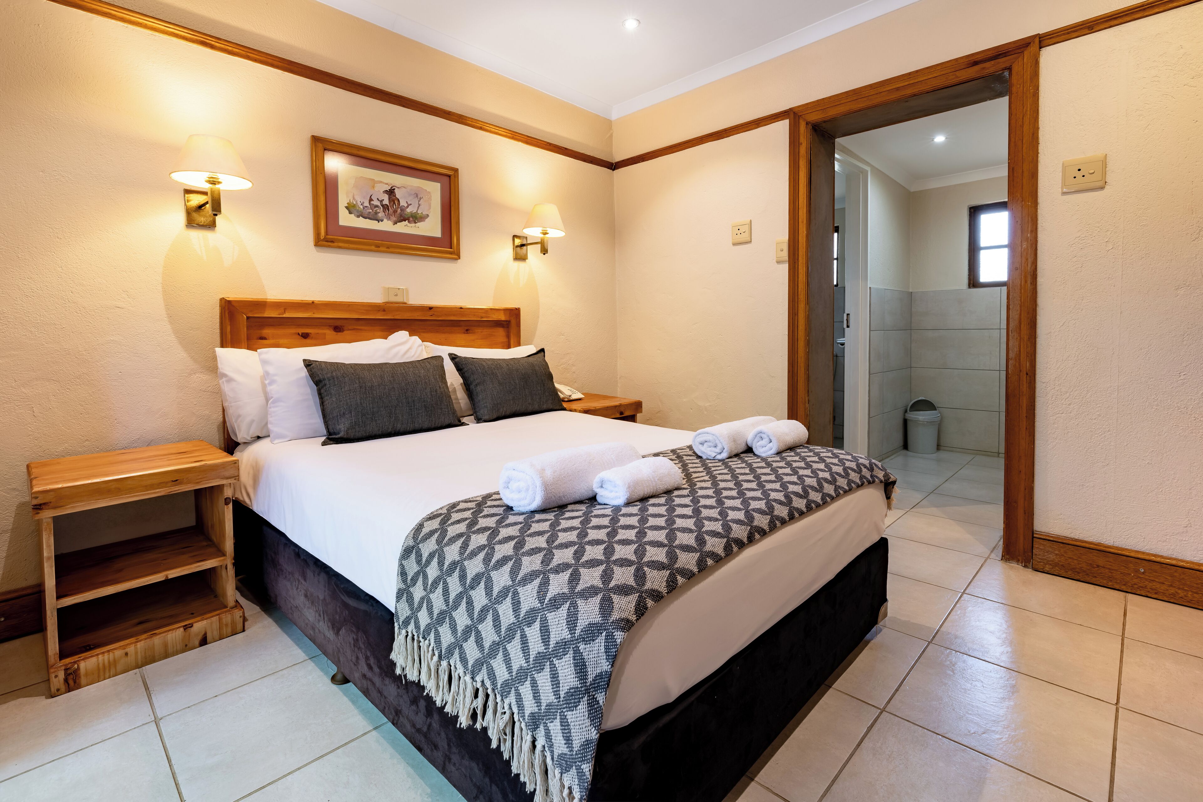A neatly made bed with a patterned throw, a wooden nightstand, and a wall-mounted lamp.