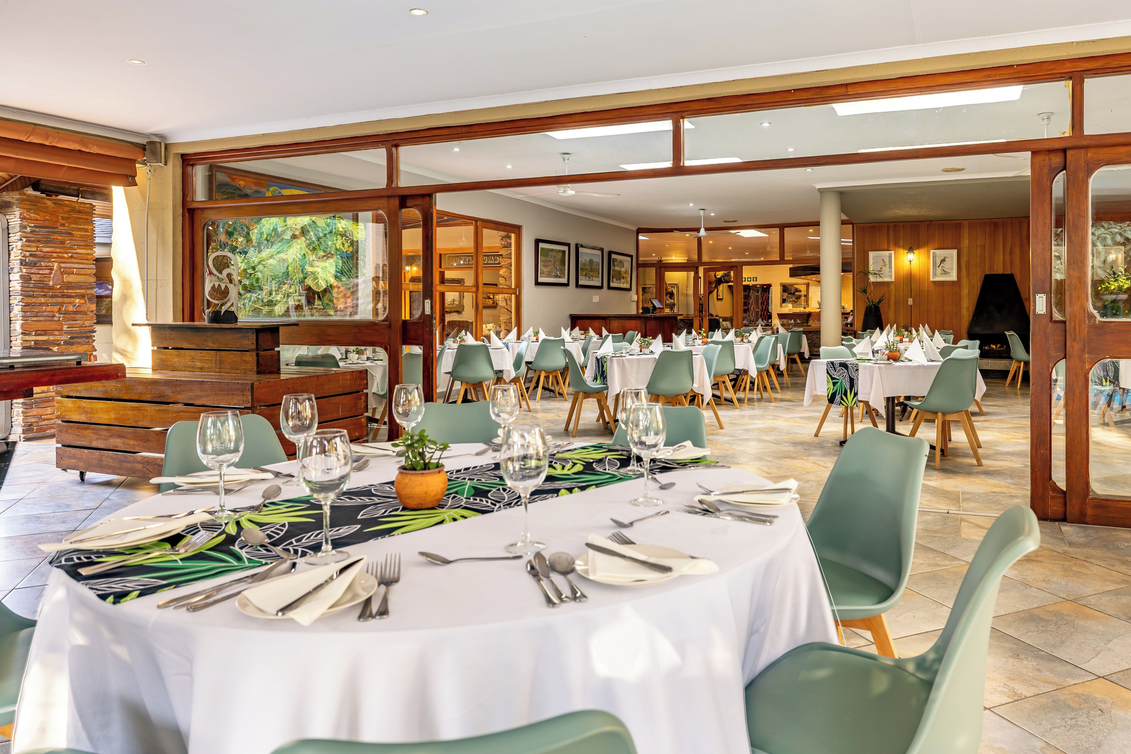 Restaurante com várias mesas preparadas para refeições, com cadeiras verdes e toalhas de mesa brancas.