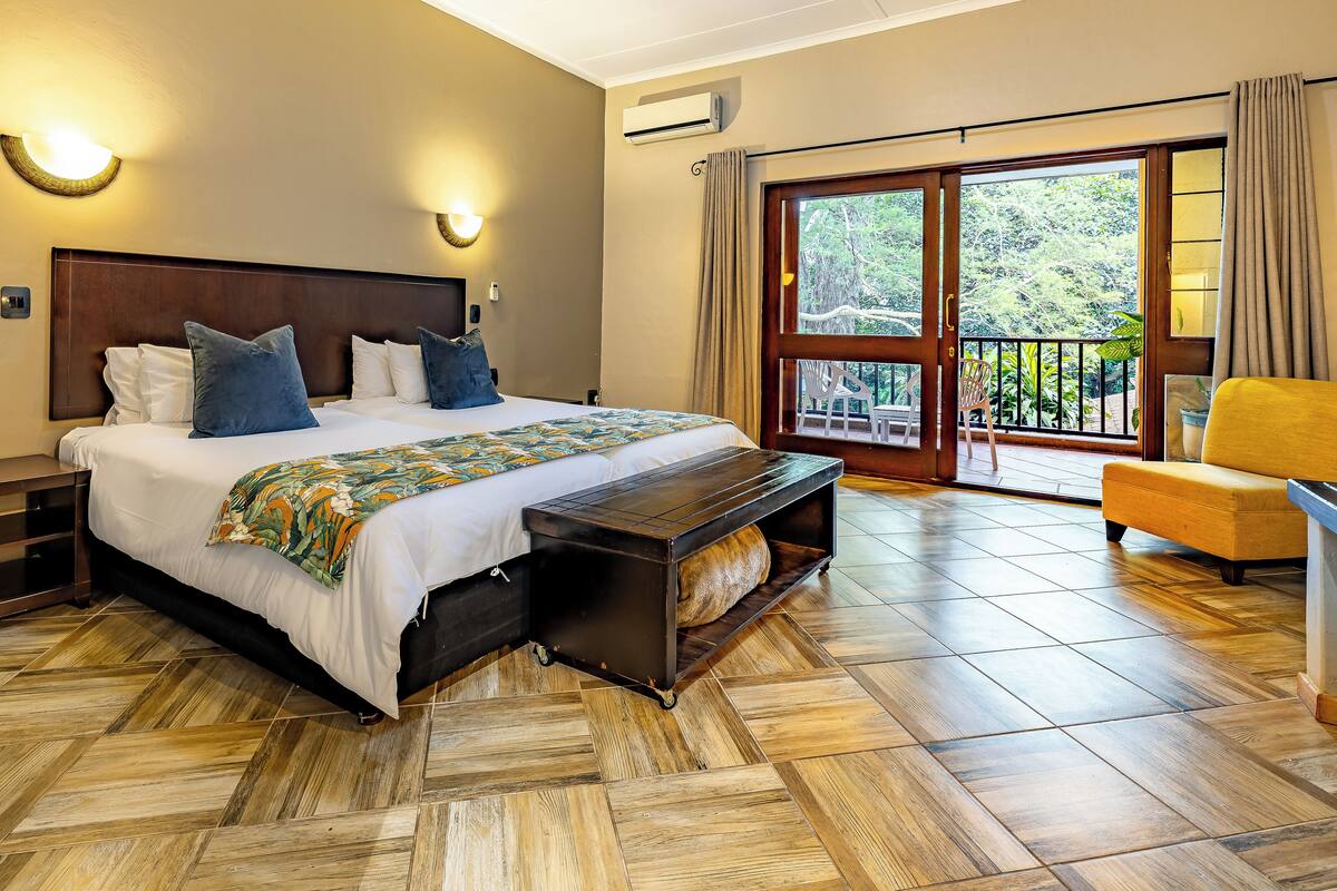 Une chambre avec un grand lit, des tables de chevet, un banc, une chaise et un balcon donnant sur un paysage verdoyant.