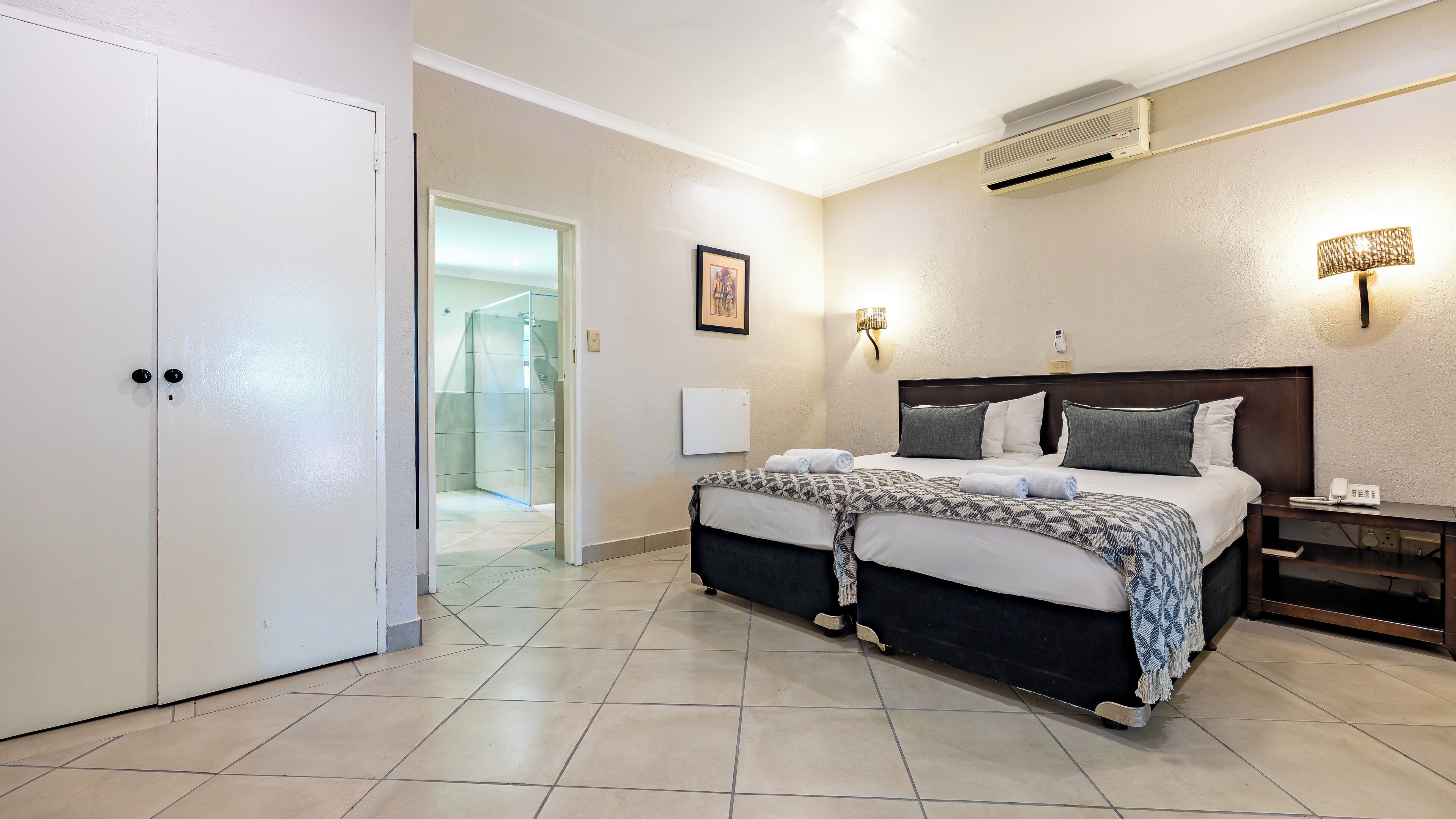 Una habitación de hotel con cama, mesita de noche, lámpara y aire acondicionado.