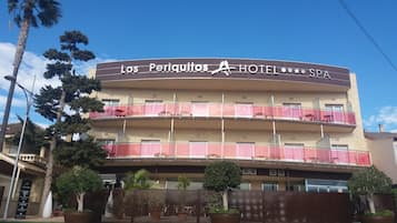 Hotellissa on parvekkeet, sen yläpuolella on kyltti, jossa lukee ”Los Periquitos Hotel SPA”, ja sen edessä on aita, jonka vieressä on ruukuissa kasveja.