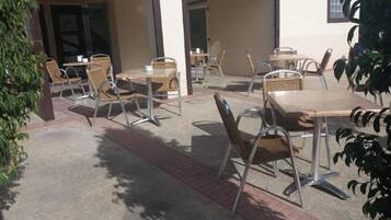 Patio esterno con diversi tavoli e sedie, un vialetto in mattoni e un edificio con facciata bianca e rivestimenti scuri.