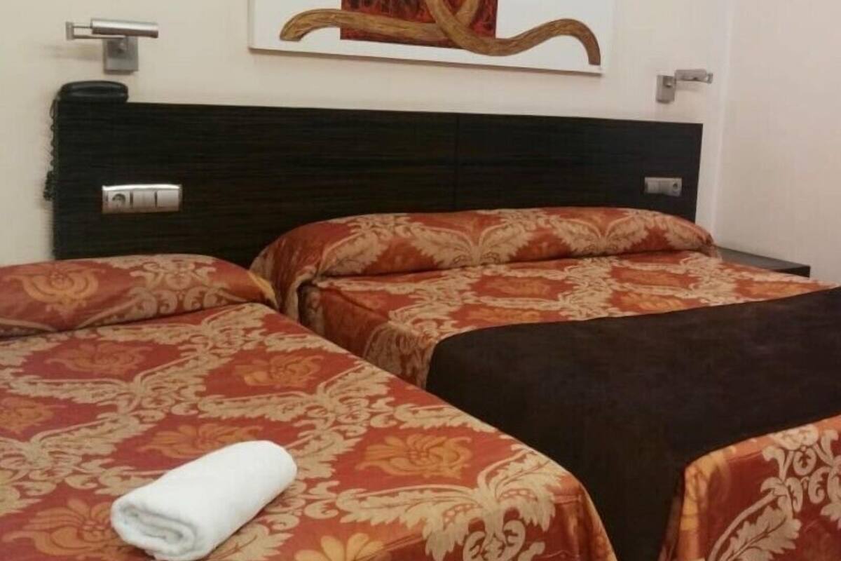A hotel room with three beds, each with a patterned bedspread and a rolled white towel on one bed.