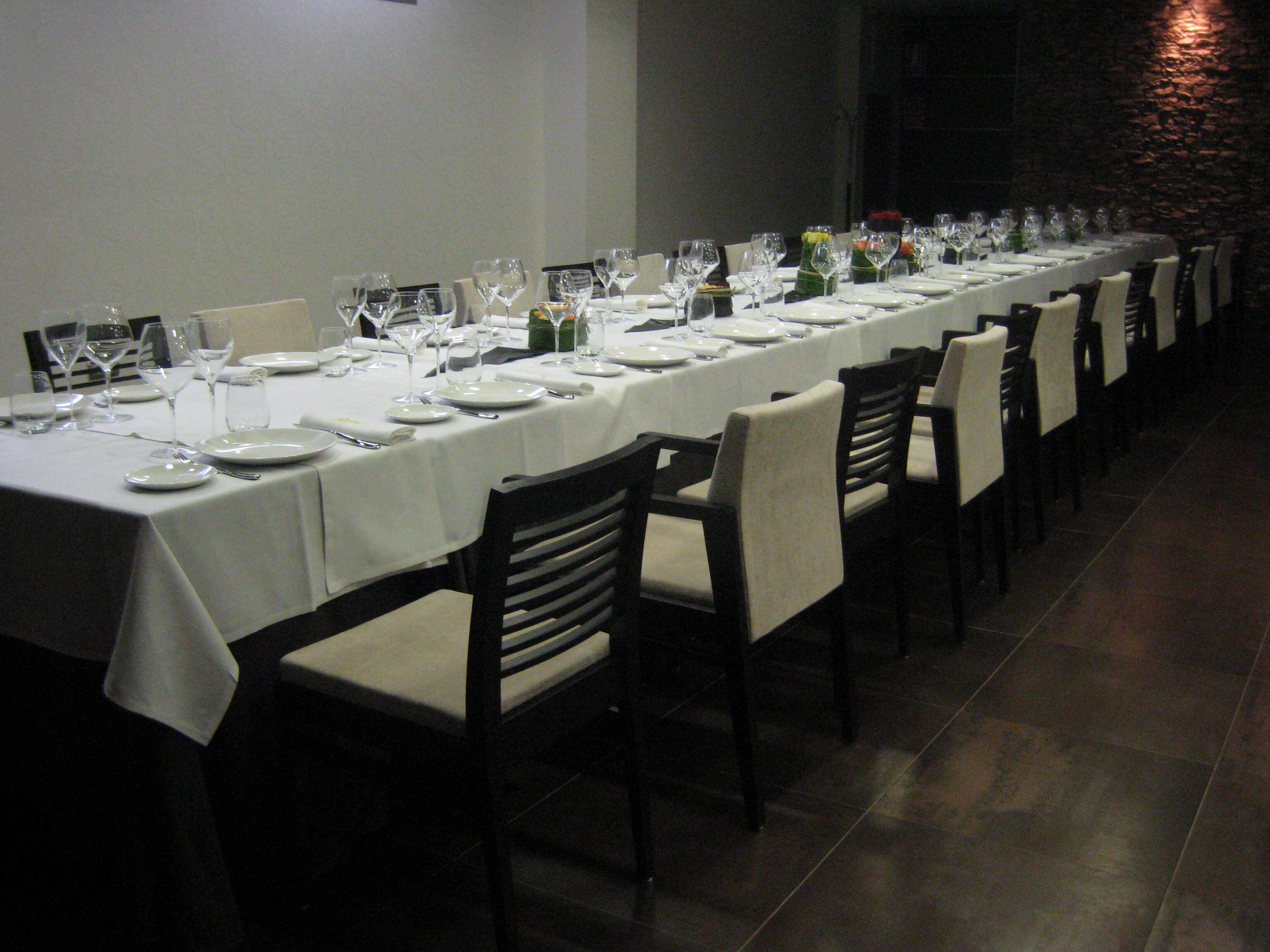 Una larga mesa de comedor con manteles blancos, platos y copas de vino, rodeada de sillas.