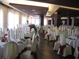 A banquet hall with rows of tables set for a formal event, featuring white tablecloths, neatly arranged chairs, and floral centerpieces.